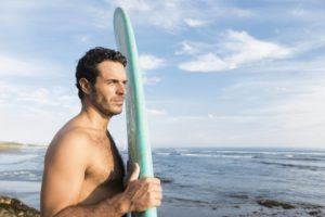 This is a color, royalty free stock photograph of a an attractive Spanish man in his 30s on a Southeast Asia beach in Bali, Indonesia. The sun is bright. He looks out at the sea water in front of him as he holds a surfboard. Photographed with a Nikon D800 DSLR camera.